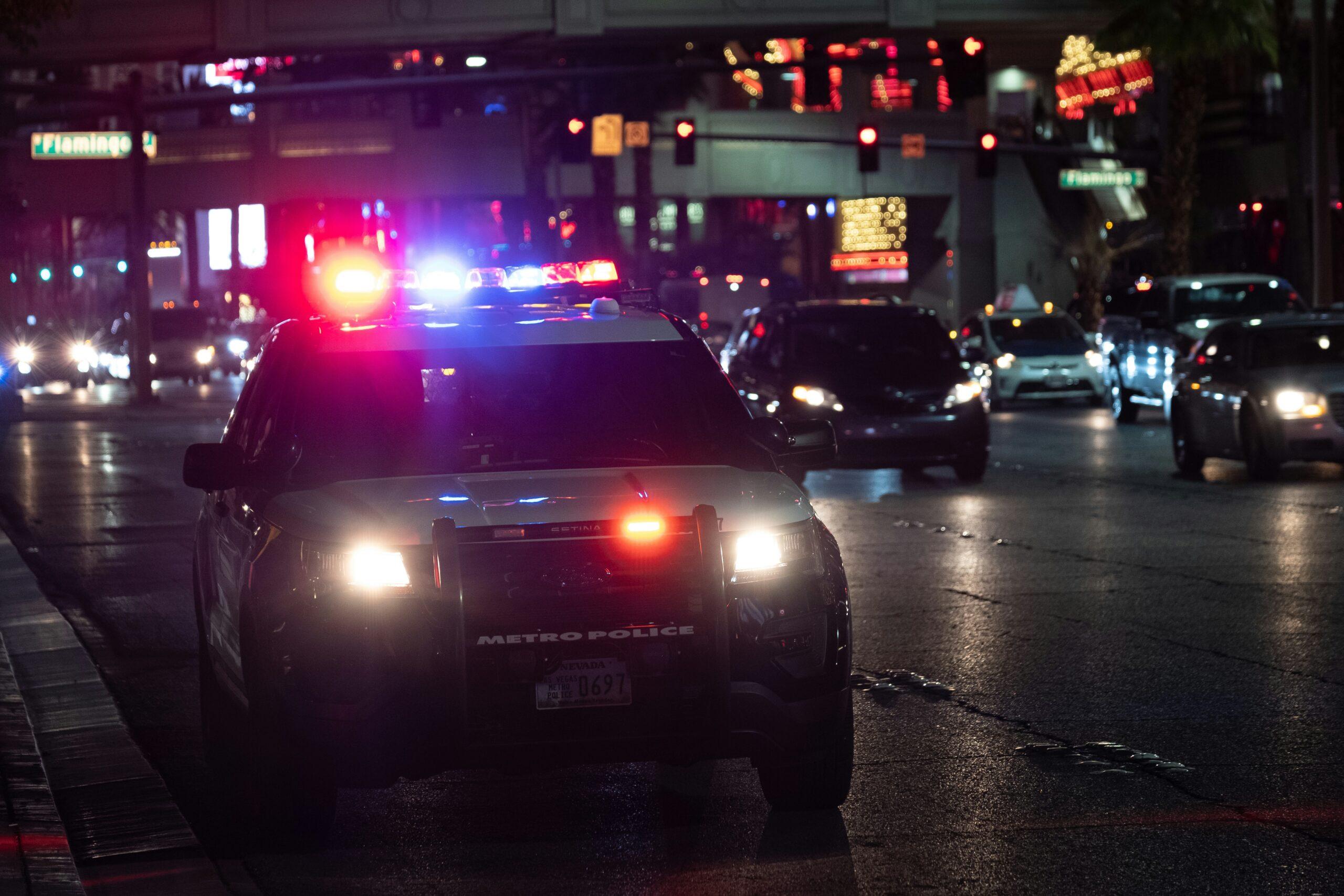 Police patrol vehicle stopped on a busy city street at night with red and blue emergency lights flashing, headlights illuminating wet asphalt, surrounded by moving traffic, traffic signals, and neon-lit buildings in the background, creating a tense urban nighttime scene with reflections on the road surface.