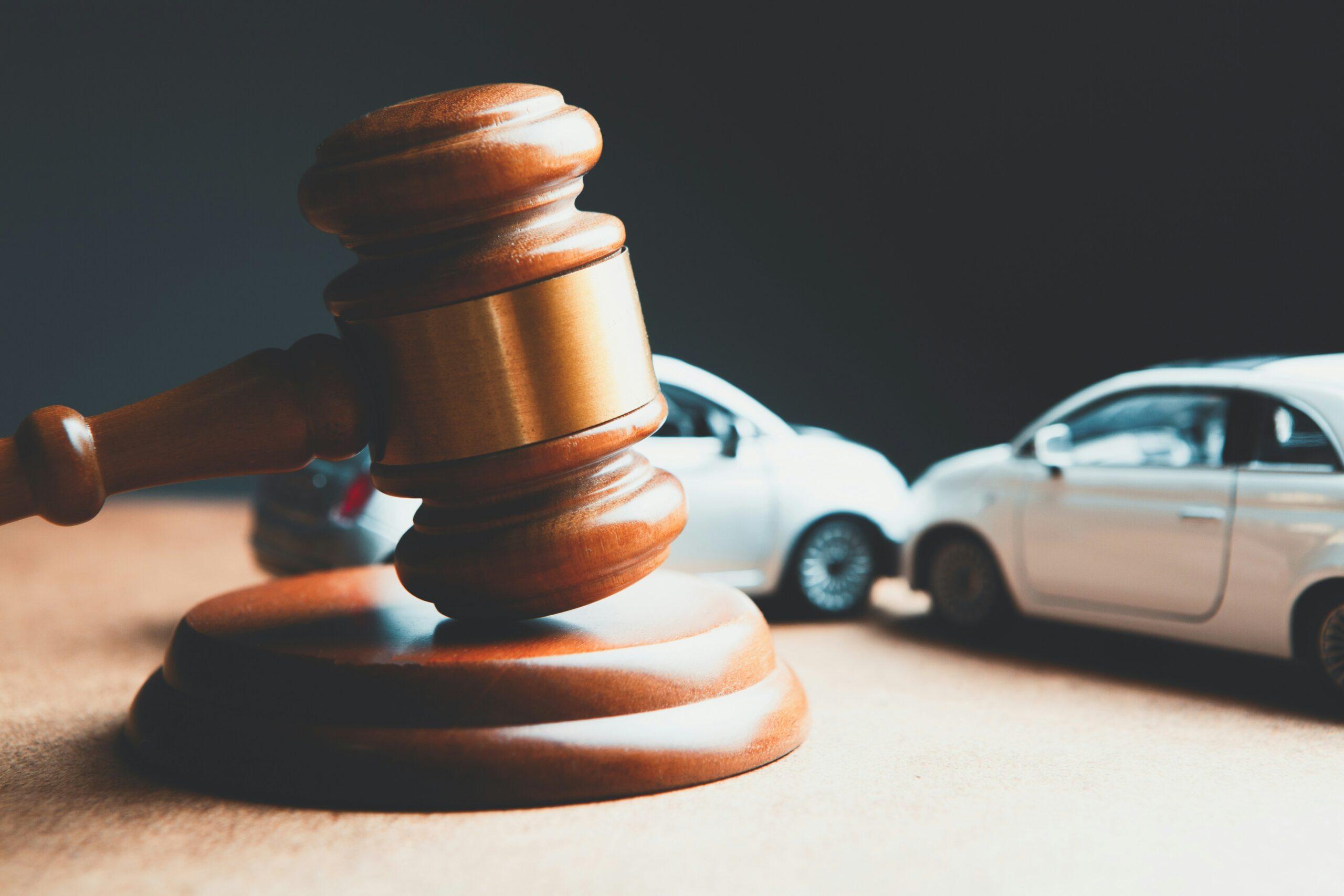 Wooden judge’s gavel resting on its sound block in the foreground, sharply lit against a dark background, with two small model cars positioned behind it facing each other as if after a collision, symbolizing a legal dispute or court decision related to a car accident or traffic incident.